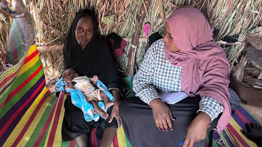Two women with a baby inside a hut made of straw, sitting on striped mat.