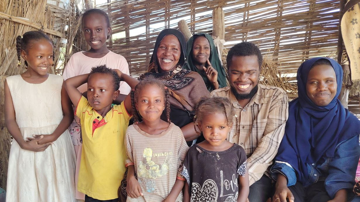 Group portrait of a large Sudanese family smiling outdoors in front of a woven wall.