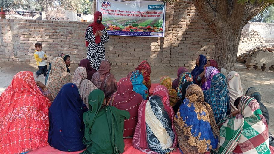 A group of women wearing colourful dresses and headscarves sit on the ground in a sunny courtyard listening to a woman teaching from the front. A sign reads ‘Provision of Tool Kits for Micro-Greenhouse Kitchen Gardens and Fruit Tree Nurseries.’