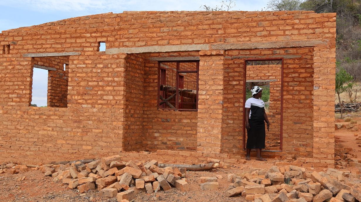 Brick house under construction with woman standing in doorway surrounded by rubble.