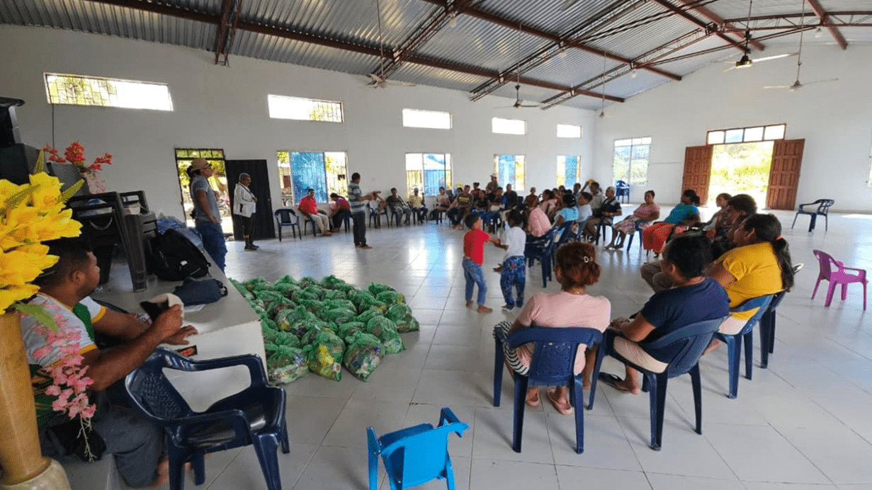 People sit in a circle inside a large hall during a community meeting, with green bags of supplies on the floor.