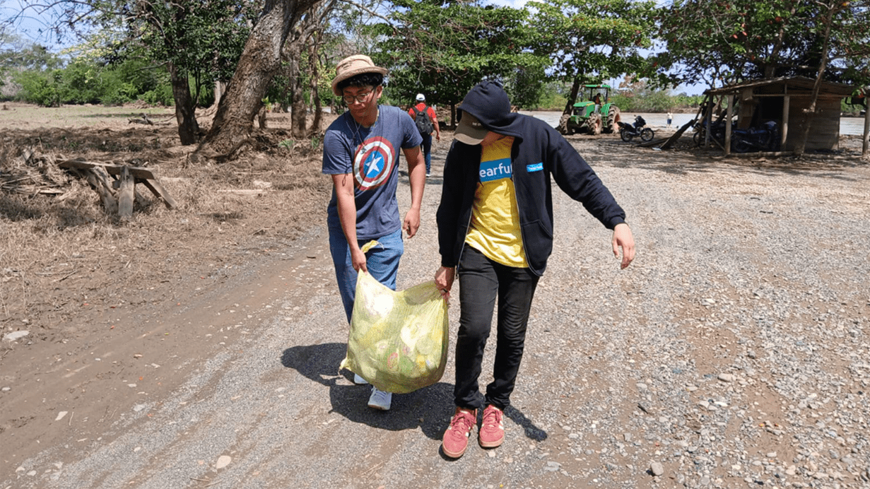 Two men carry a large bag of supplies along a gravel road near trees and small buildings.