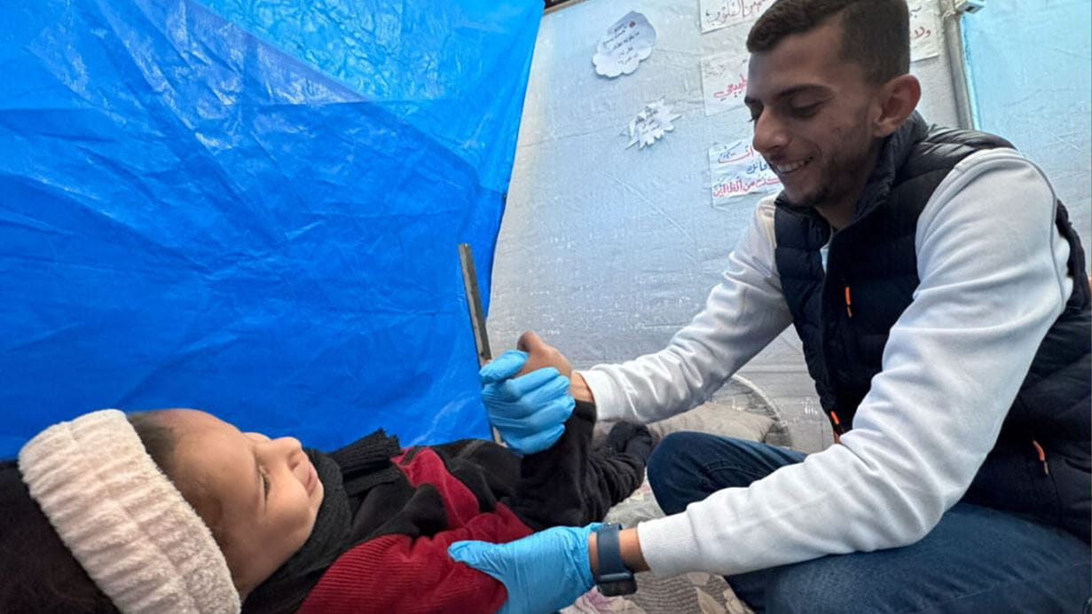 A child lays on the ground and a medical worker wearing blue gloves holds a her arm and hand, they both smile.