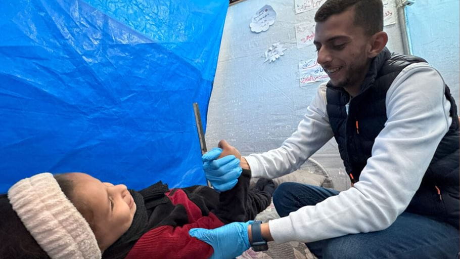 A child lays on the ground and a medical worker wearing blue gloves holds a her arm and hand, they both smile.