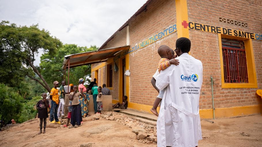 A healthcare worker carries a child in front of a rural health center in Africa.