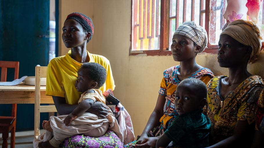 Three African women sit with their children, all looking towards the right side of the frame.