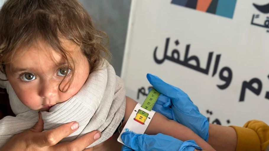 Health worker measures a child's arm with a measuring tape, Arabic text on background.