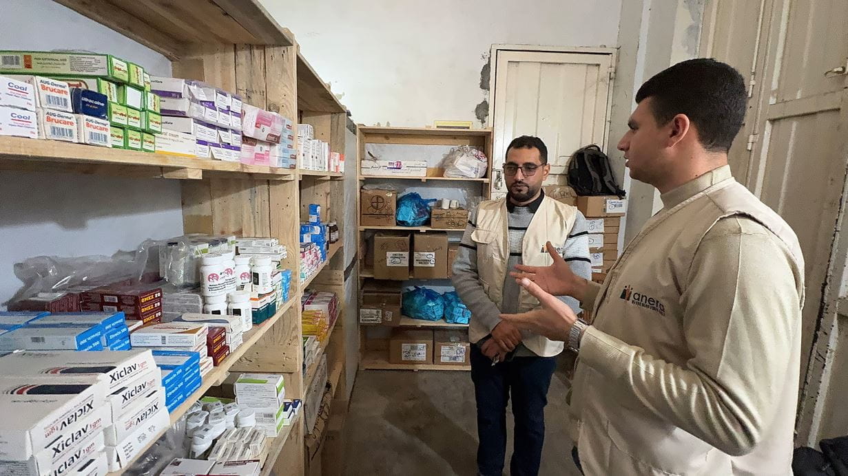 Two men in a storage room filled with shelves of medical supplies and medications.