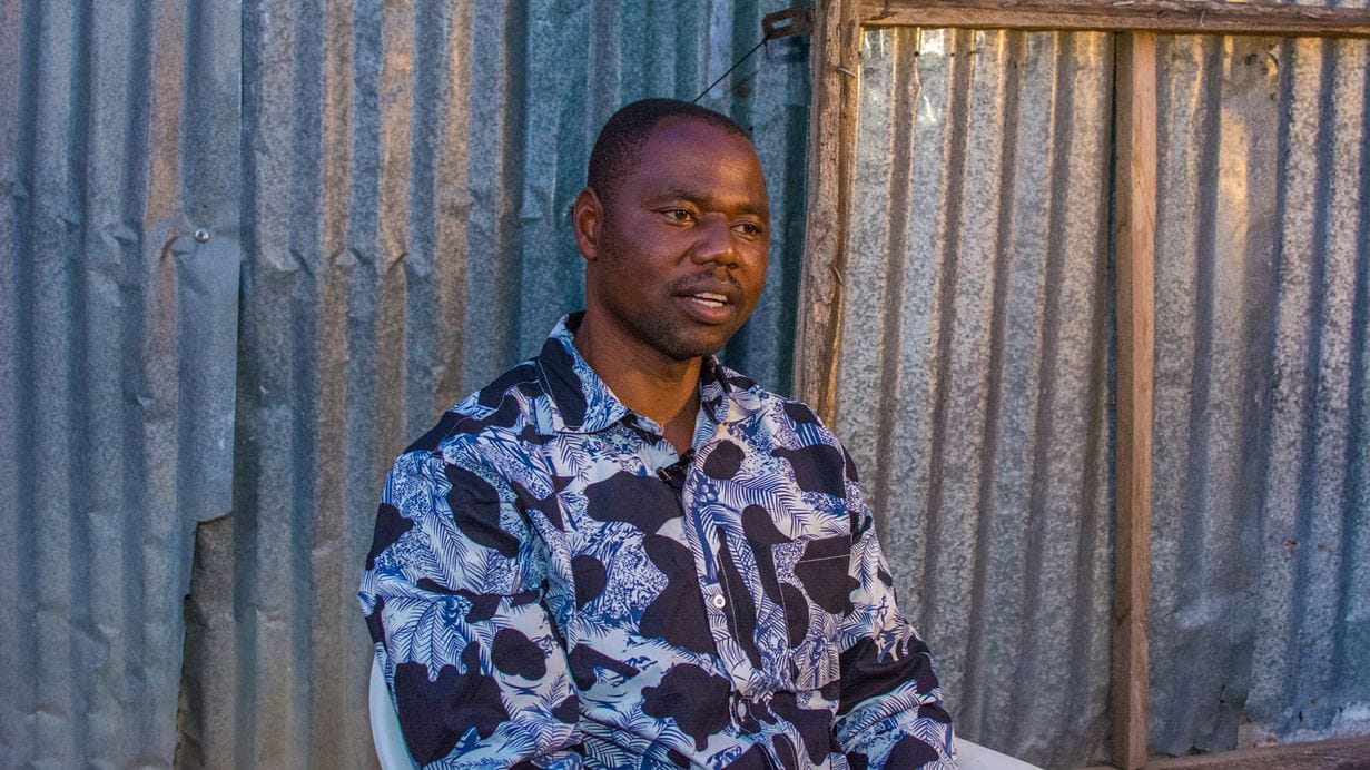 Rev Canon Chukle, the church leader from Fwagul, Nigeria. He’s wearing a blue and white patterned shirt, and sits in front of a corrugated metal wall during an interview.