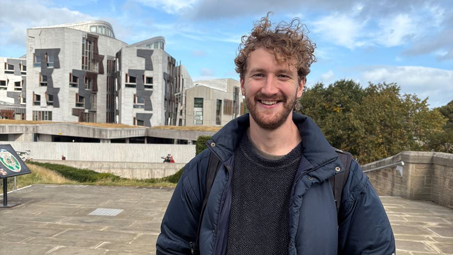 Smiling man with curly hair standing in front of the Scottish Parliament building.