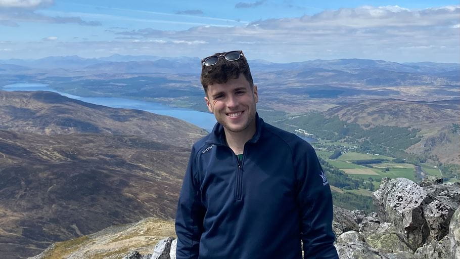 Man smiling on a mountain peak with lakes and hills visible in the background.