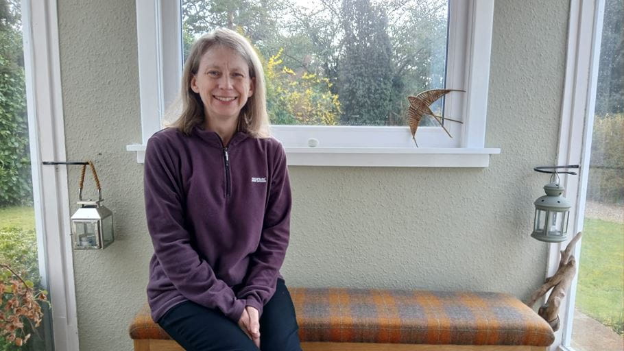 A woman sits on a bench smiling at the camera in a room with windows looking out to a garden.