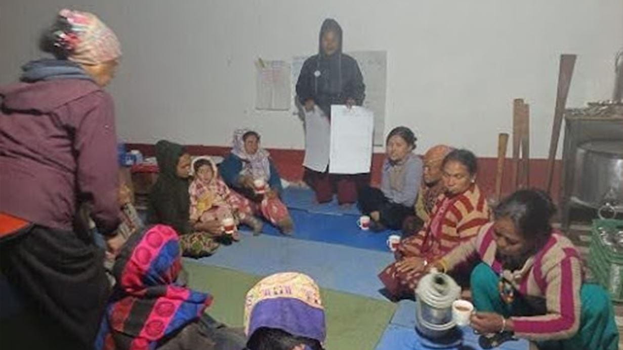 A group of women sit on floor for an indoor meeting, with one woman holding papers standing.