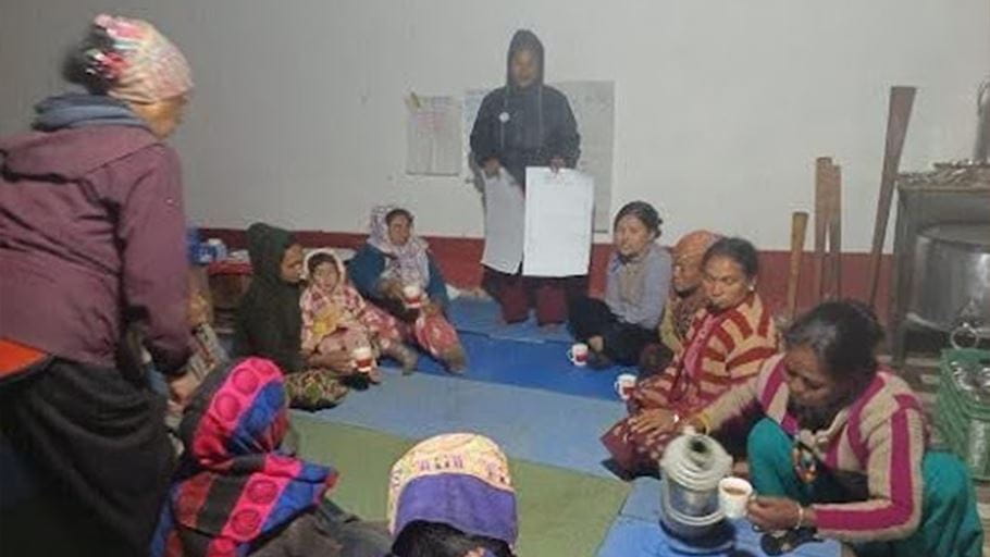A group of women sit on floor for an indoor meeting, with one woman holding papers standing.