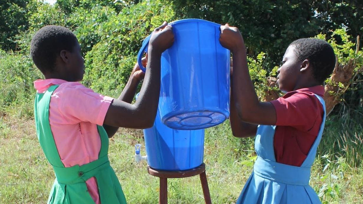 Two schoolgirls lifting a blue bucket, possibly filled with water, outdoors. Handwashing.