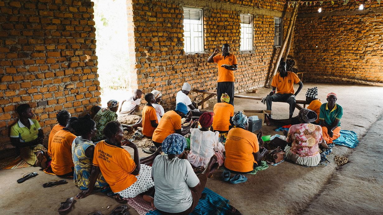 A group of adults, mostly women, sit on the floor in a brick-walled room, wearing orange shirts and listening to a person standing and speaking.