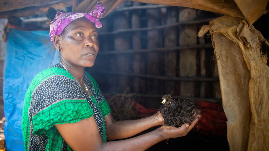 Woman holds soil in hands in front of structure, hopeful expression, pink headscarf.