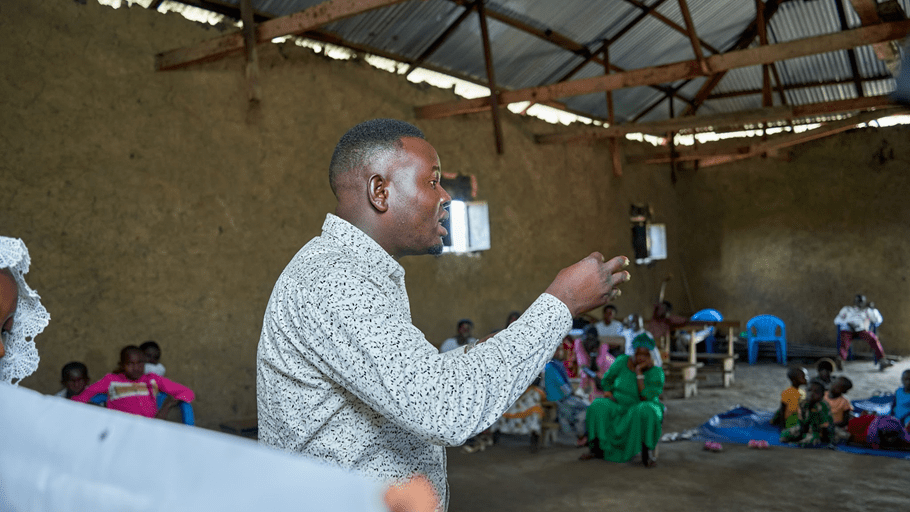 A man in a patterned shirt gestures while speaking to a group of people seated in a room with dirt walls and a corrugated metal roof.