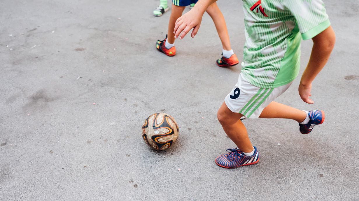 Child in green and white jersey dribbling a worn soccer ball on a concrete surface.