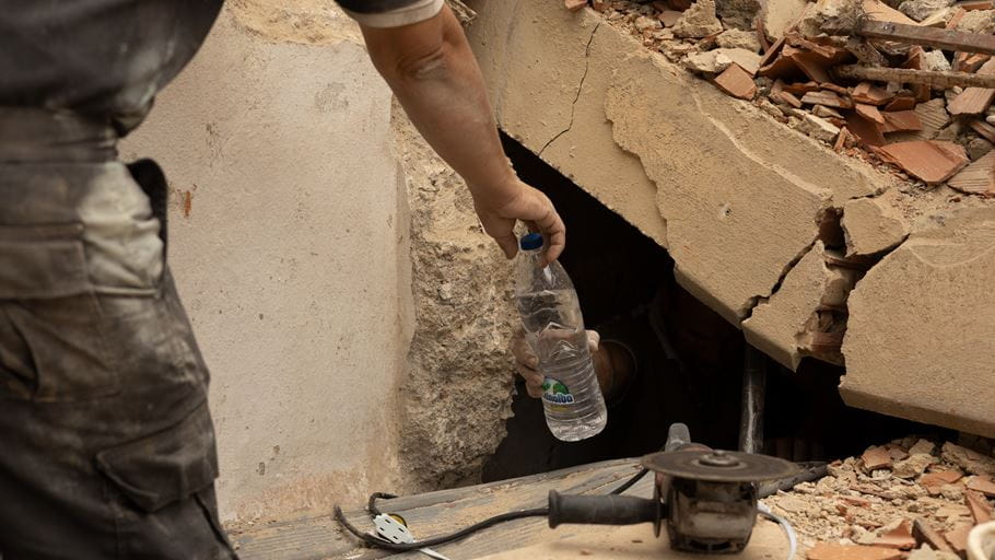 A hand offers a water bottle into a narrow opening in concrete rubble, implying a rescue.
