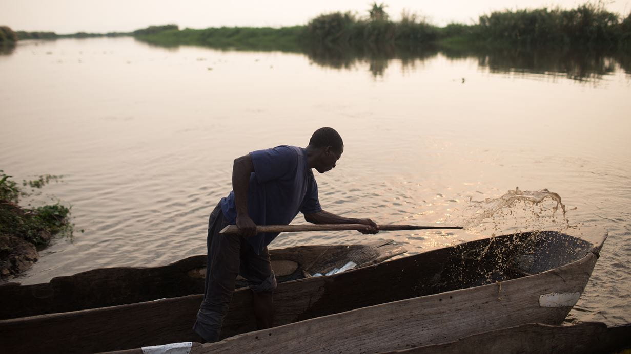 A man in a blue shirt and dark pants is paddling a wooden canoe in a calm river in Mozambique at sunset, with lush greenery visible along the banks.