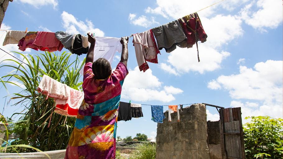 A woman in a colorful dress hangs laundry on a clothesline under a bright blue sky with scattered clouds, surrounded by greenery and a stone wall in the background.