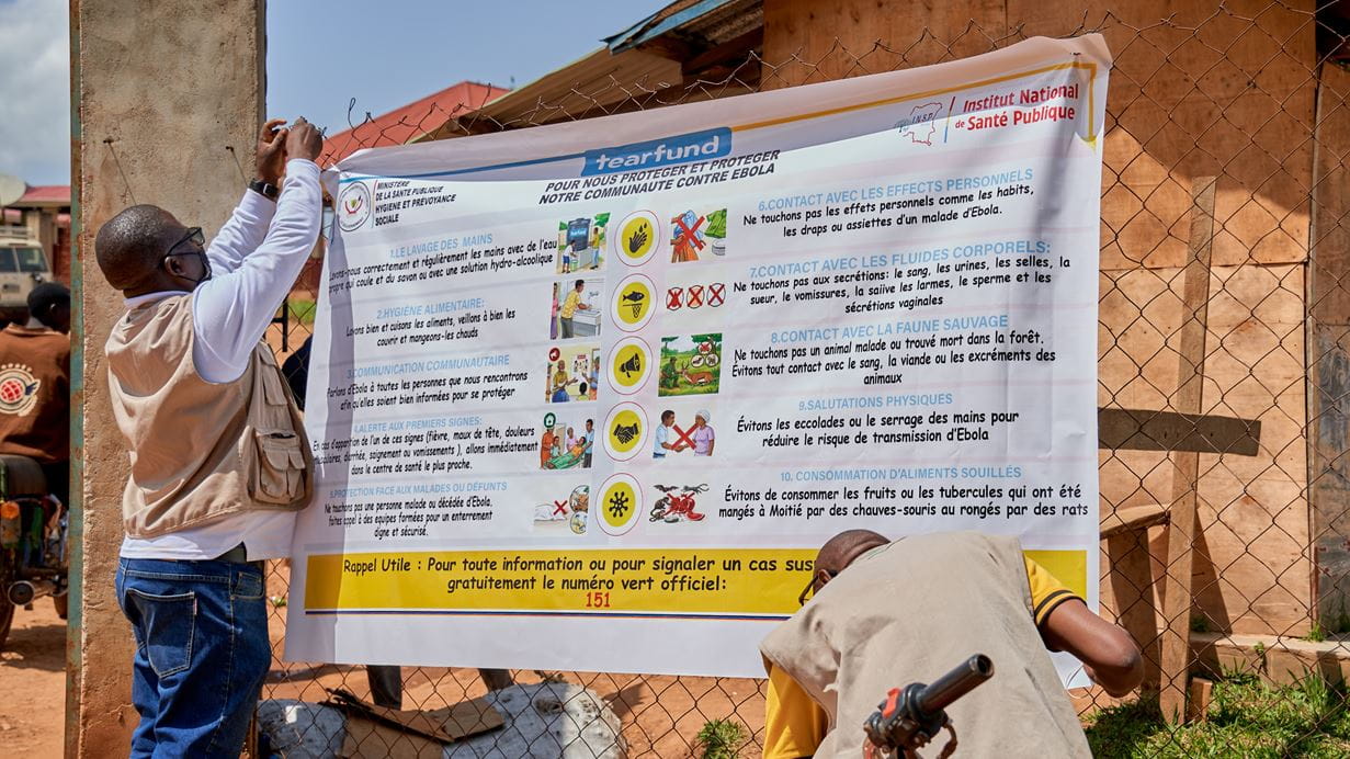 Two men hang a large French banner detailing Ebola prevention measures outdoors.