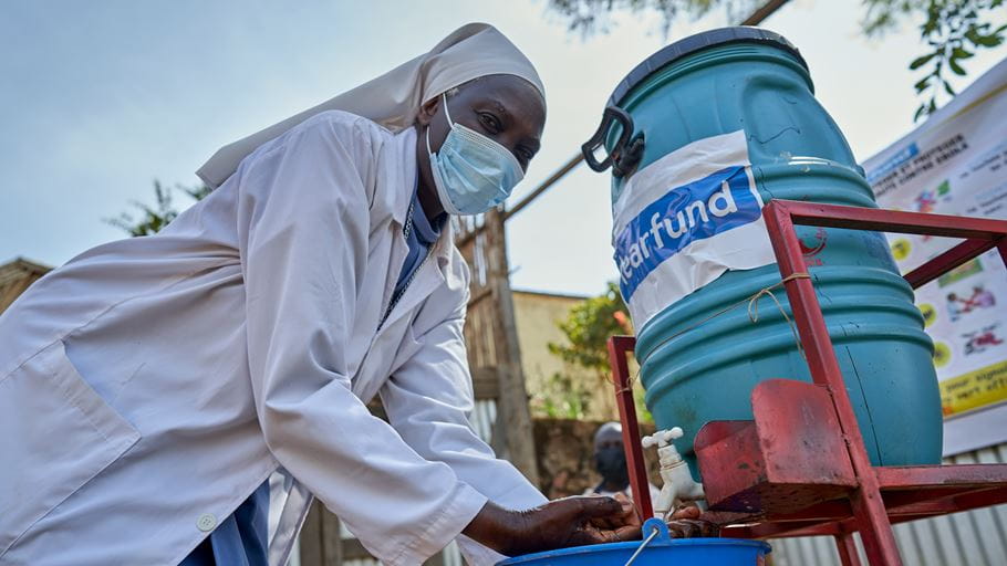 Woman in mask and lab coat washing hands at an outdoor handwashing station with a blue barrel.