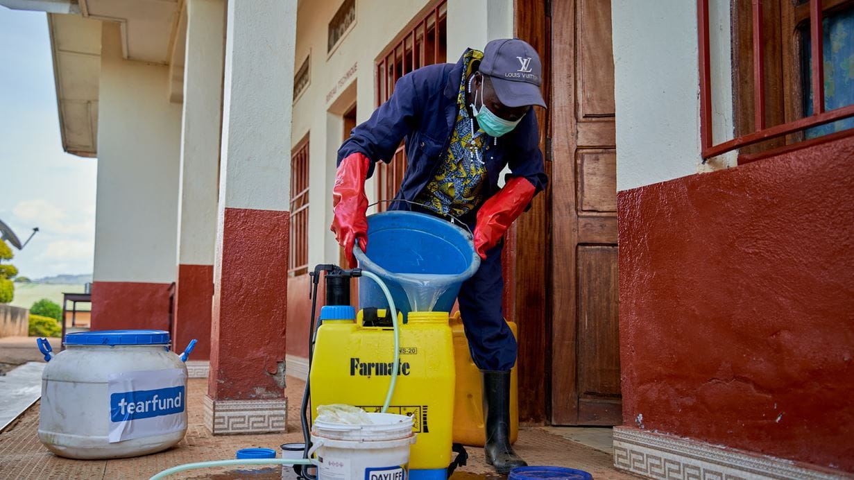 Person in PPE pouring liquid into a sprayer on a porch, preparing for disinfection.