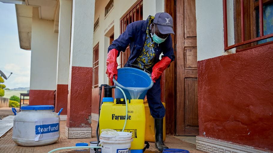 Person in PPE pouring liquid into a sprayer on a porch, preparing for disinfection.