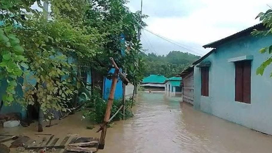 Village street heavily flooded with muddy water, surrounded by houses and lush green trees.