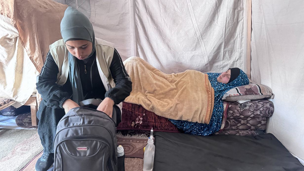 A woman in a headscarf packs a backpack inside a tent; another person rests on a bed.