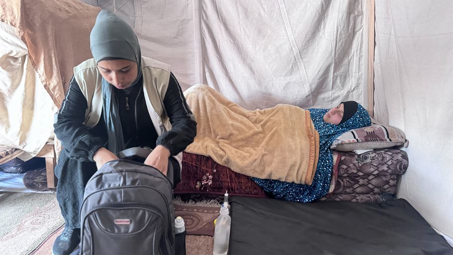 A woman in a headscarf packs a backpack inside a tent; another person rests on a bed.