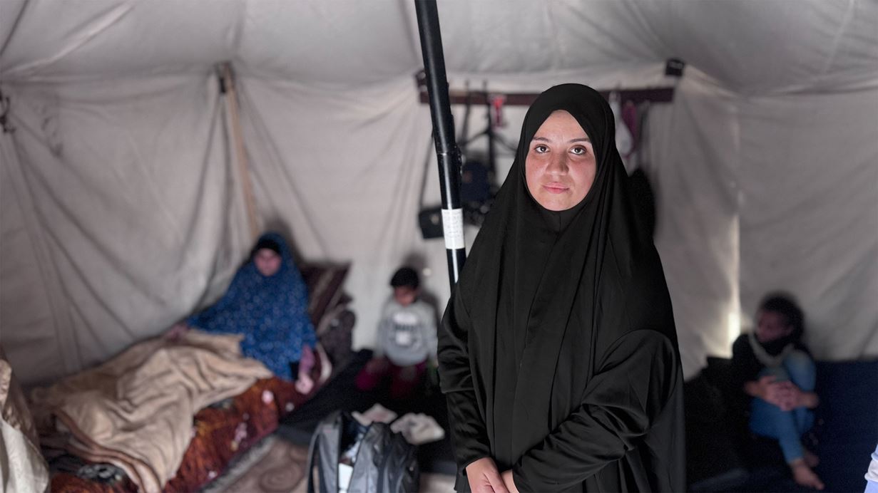 Woman in black hijab stands in a tent, with family members in the blurry background.