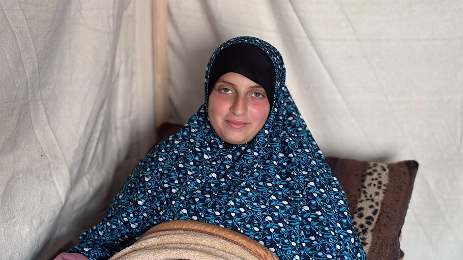 A woman wearing a blue patterned hijab smiles from inside a tent, covered by a blanket.
