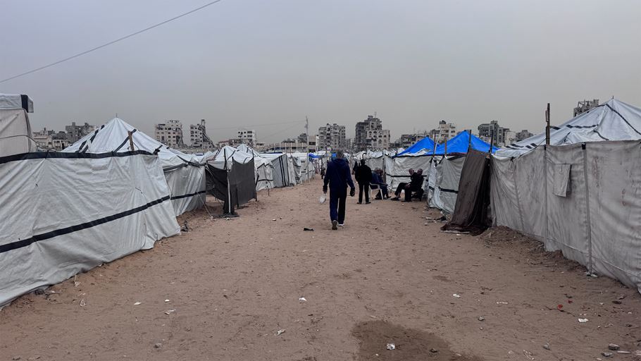 Makeshift refugee camp with rows of grey and blue tents, people, and distant damaged buildings.