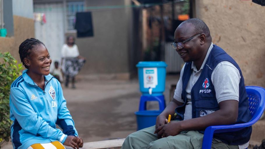 A smiling woman in a light blue tracksuit talks to a smiling man in glasses and a blue PPSSP vest. They are outdoors, with a blue water dispenser and residential buildings in the background.