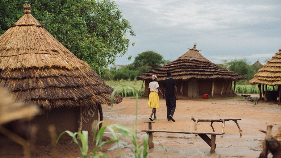 A man and woman walk away from the viewer on a muddy path, past several traditional round huts with thatched roofs, under an overcast sky.