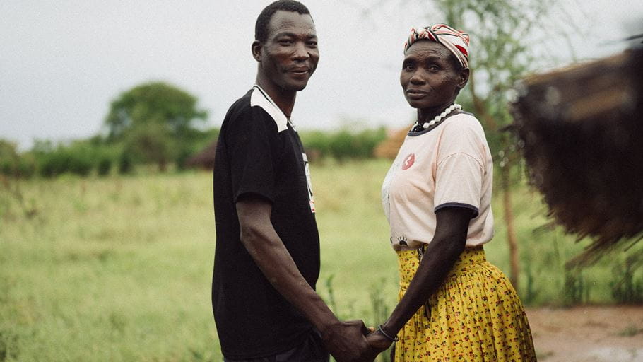 A man and a woman stand holding hands outdoors. The man wears a black t-shirt and the woman a pink t-shirt, yellow floral skirt, and striped headscarf. They both look towards the viewer with a grassy field in the background.
