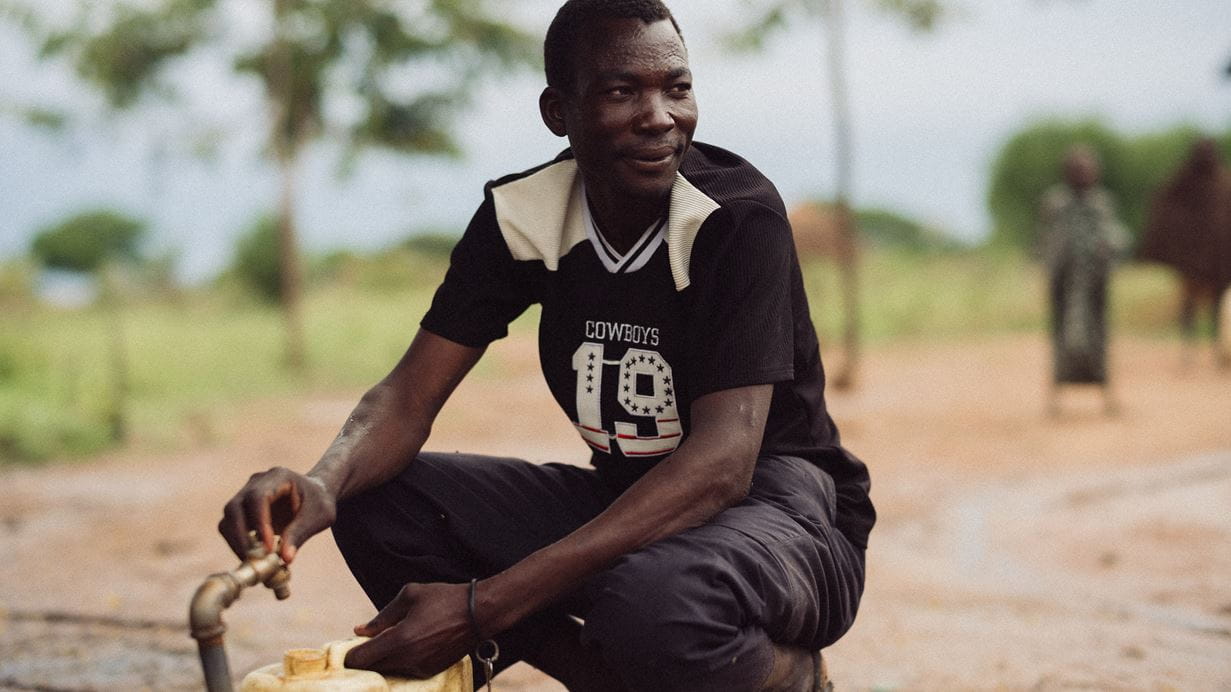 A man in a black 'Cowboys 19' t-shirt crouches, operating a brass tap to fill a yellow jerrycan. He looks to his right with a gentle smile in an outdoor setting.
