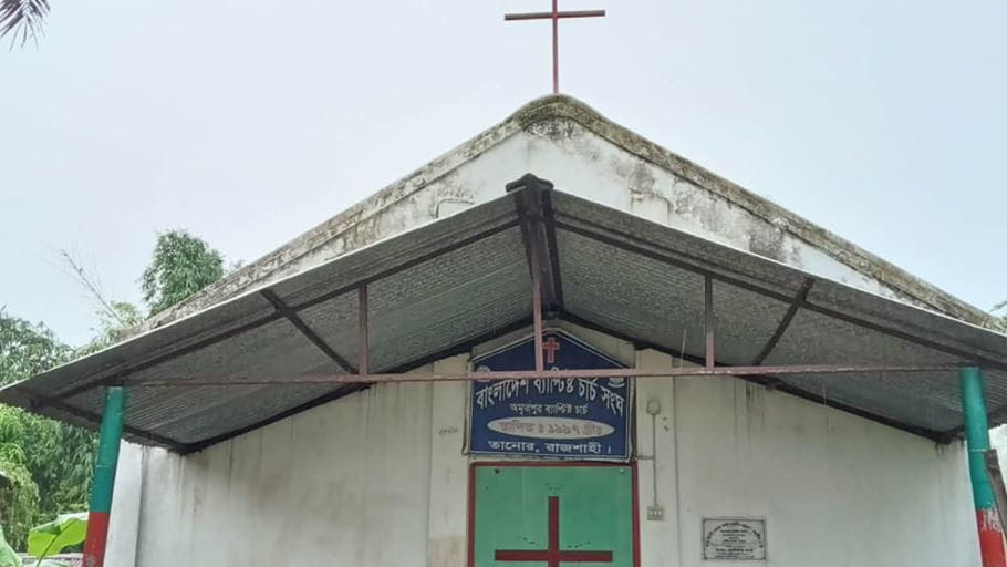 Front view of a simple church building with white walls, a corrugated metal canopy, and a red cross on its roof. A blue sign with Bengali text is visible above the green double doors, which also feature a red cross.