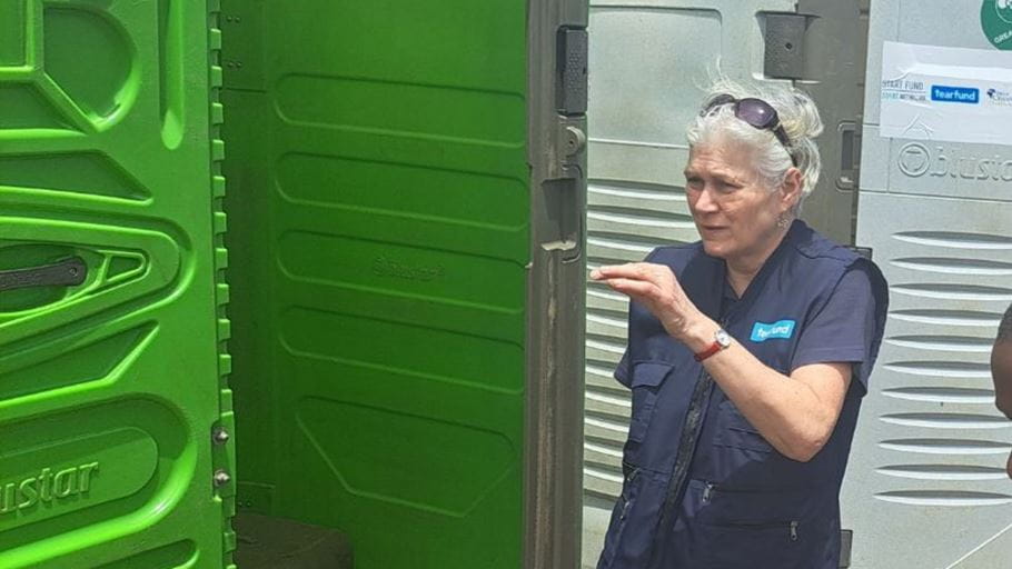A woman with grey hair and sunglasses on her head, wearing a blue vest, gestures with her right hand. She stands between an open green portable toilet and a grey portable toilet with a 'Tearfund' sticker.