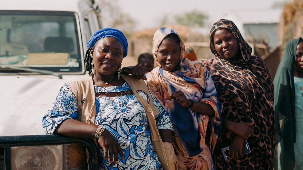 Three women, two smiling, stand outdoors in front of a white vehicle. The woman on the left wears a blue headscarf and patterned dress, leaning on the car's bumper. The central woman wears a colourful wrap and leans on the first. The woman on the right wears a leopard print headscarf.