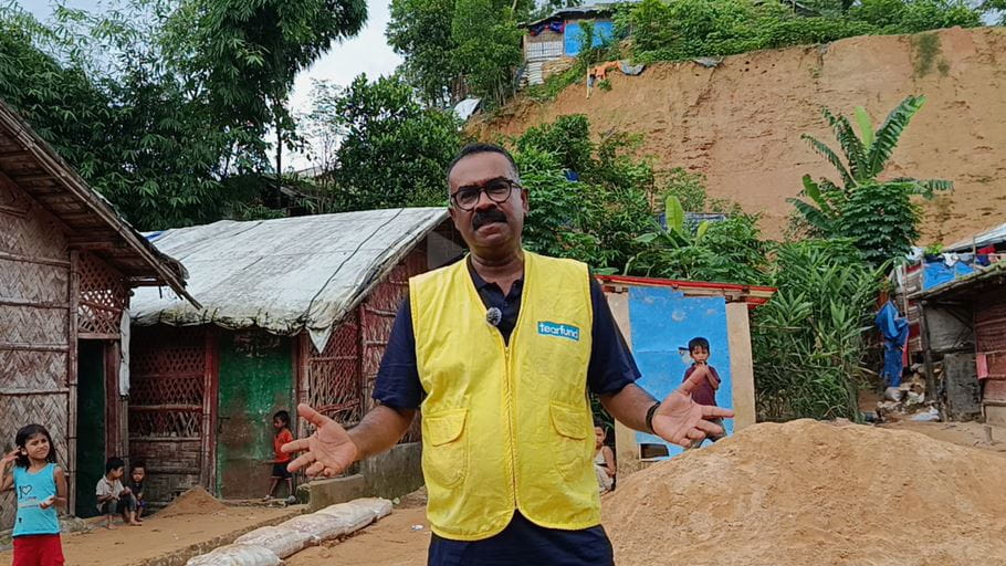 A man in a yellow Tearfund vest gestures with open hands in front of basic shelters and children, with a steep, bare earth hillside behind him.