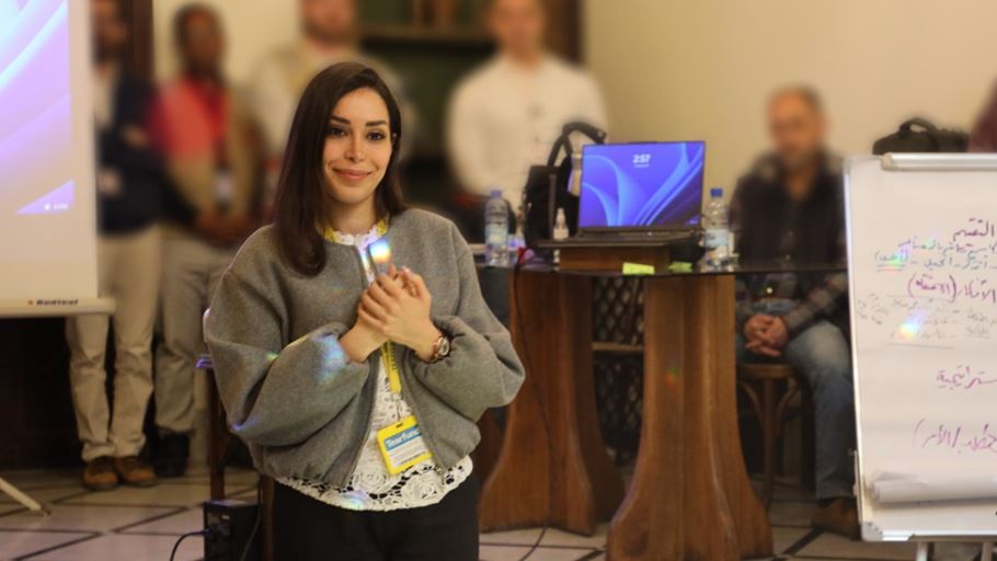 A smiling woman wearing a grey jacket and yellow lanyard holds a glowing rectangular device. Blurred people, a projector screen, a laptop, and a whiteboard are in the background.