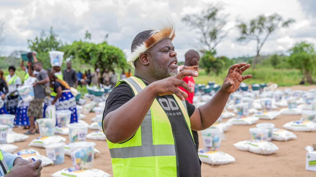 A man with a grey beard and a traditional feathered headpiece, wearing a black t-shirt and a bright yellow high-visibility vest, gestures with both hands. In the background, people are gathered around numerous white sacks and clear buckets arranged on the bare ground.