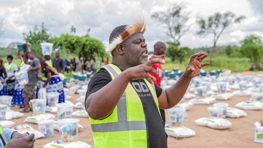 A man with a grey beard and a traditional feathered headpiece, wearing a black t-shirt and a bright yellow high-visibility vest, gestures with both hands. In the background, people are gathered around numerous white sacks and clear buckets arranged on the bare ground.