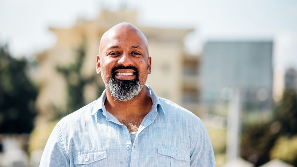 Bald man with a grey beard smiling broadly, wearing a light blue shirt, in an outdoor urban setting.