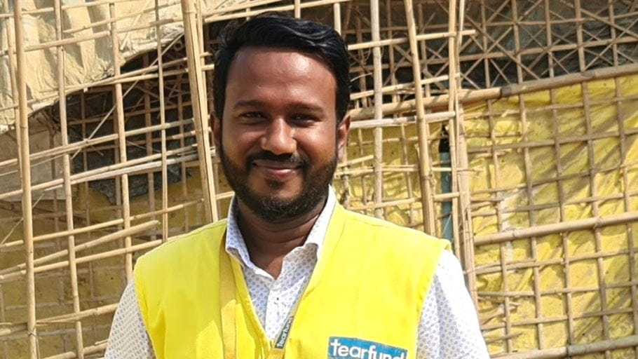 Smiling man with a beard wearing a yellow Tearfund vest in front of a bamboo structure.