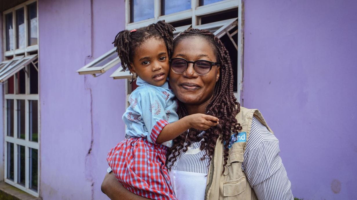 A woman with dreadlocks and glasses holds a young girl with pigtails. Both smile warmly in front of a vibrant purple building.
